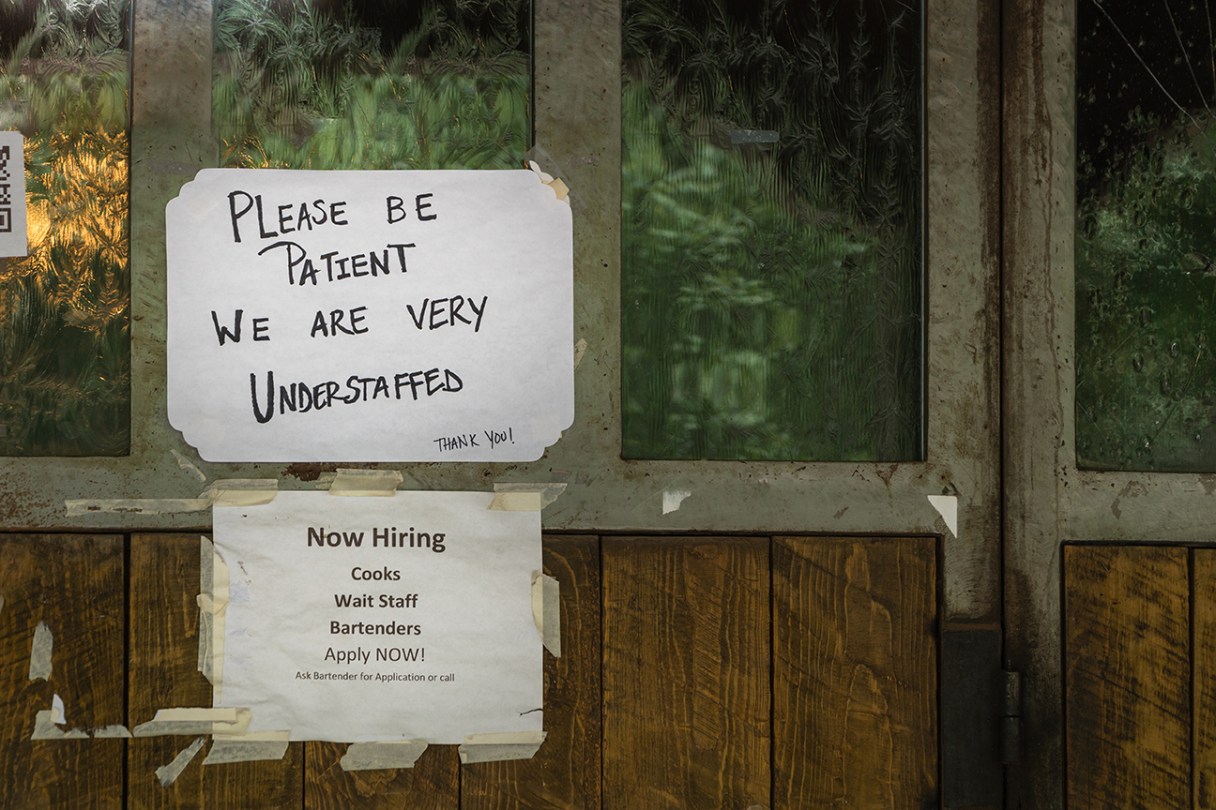 Rustic sign in restaurant window during time of Covid stating restaurant is understaffed and is hiring in a struggling economy, showing the plight of small businesses in the post-pandemic economy.